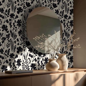 May include: Interior shot with black and cream floral wallpaper. An oval mirror reflects the room. Two beige vases with dried flowers sit on a light wood cabinet. The scene is a study in contrasts.