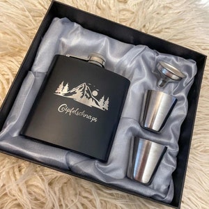 Könnte beinhalten: Eine schwarze Trinkflasche mit einem Berg- und Baummotiv und dem Text "Gipfel Schnaps". Die Flasche befindet sich in einer schwarzen Box mit silberner Auskleidung. Die Box enthält außerdem zwei silberne Schnapsgläser und einen silbernen Trichter.