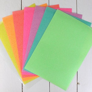 Puede incluir: Un conjunto de ocho hojas de papel de purpurina en varios colores, incluyendo blanco, amarillo, naranja, rosa, morado, verde azulado y verde. Las hojas est&aacute;n dispuestas en forma de abanico, con la hoja verde en la parte superior.