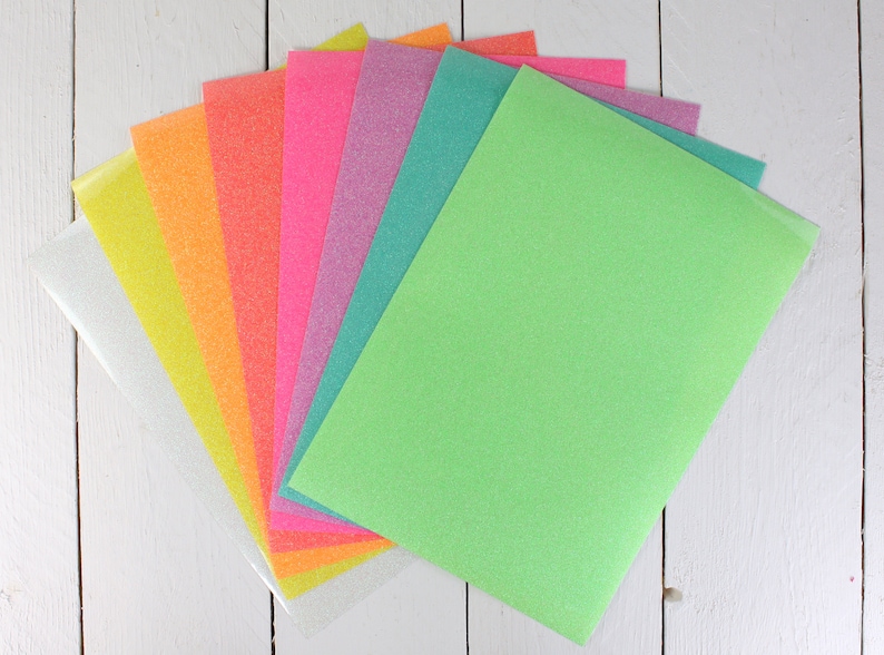 Puede incluir: Un conjunto de ocho hojas de papel de purpurina en varios colores, incluyendo blanco, amarillo, naranja, rosa, morado, verde azulado y verde. Las hojas est&aacute;n dispuestas en forma de abanico, con la hoja verde en la parte superior.