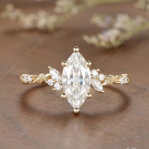 May include: A gold ring featuring a central marquise-cut clear gemstone held by six prongs. The band is adorned with smaller marquise and round-cut clear stones arranged in a cluster on both sides, set in a polished gold metal band.
