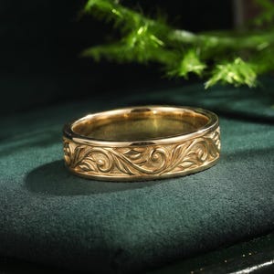 May include: A gold ring featuring an intricate, raised scrollwork pattern that wraps around the entire outer band. The metal has a polished finish, highlighting the detailed swirling leaf and vine engravings across the surface of the band.