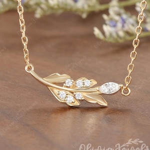 May include: A gold-colored necklace featuring a leaf design with embedded clear gemstones. The leaf is positioned horizontally, with the chain extending from both ends. The necklace is set against a blurred background.