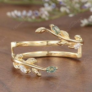 Könnte beinhalten: Ein goldener Bypass-Ring mit zwei zarten Zweigen und strukturierten Blättern. Der Ring ist mit zwei marquise-geschliffenen grünen Moosachat-Steinen und zwei klaren marquise-geschliffenen Steinen besetzt, was ein naturinspiriertes Design ergibt.
