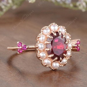May include: A rose gold ring with a large red gemstone surrounded by a halo of white pearls. The ring has a small pink gemstone arrow on each side of the main stone.
