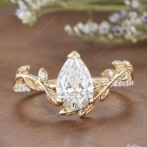 May include: A pear-shaped diamond engagement ring with a delicate gold band and leaf-shaped accents. The ring is set on a wooden surface.