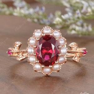May include: A rose gold ring with a large oval ruby gemstone surrounded by a halo of white pearls. The ring has two small ruby accents on the sides.
