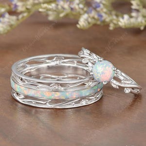 May include: A set of two silver rings. One ring features a large, round opal stone set in a leaf design. The other ring has a band with an opal inlay and a delicate, openwork design. The rings are set against a blurred floral background.