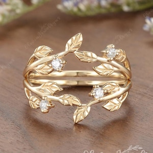 May include: A gold ring featuring a leaf design and small, clear gemstones. The ring has a double band and is decorated with detailed leaf and branch motifs. The ring is on a wooden surface.