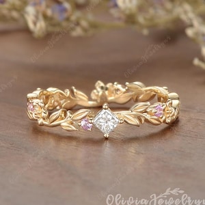 May include: A gold ring featuring a central princess-cut clear stone flanked by small pink round stones. The band is designed with a continuous pattern of sculpted leaves and vines that wrap around the entire circumference of the jewelry piece.