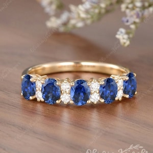 May include: A gold band ring featuring a series of oval-cut, deep blue gemstones alternating with smaller, round, clear gemstones. The ring is set against a blurred background of wood and flowers.