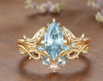 May include: A gold ring featuring a central marquise-cut light blue gemstone, flanked by two dolphin motifs. The band is accented with smaller round light blue stones and a single white pearl at the base, set within a decorative metal framework.