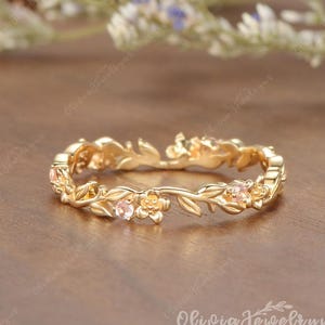 May include: A gold ring with a floral design. The ring features delicate leaves, small flowers, and several small, light pink gemstones. The ring is set on a wooden surface with blurred floral elements in the background.