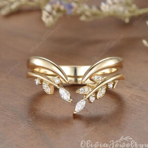 May include: A gold ring featuring a V-shaped band with two symmetrical leaf-like branches extending from the center. Each branch is adorned with small, clear, round-cut stones and a single marquise-cut stone at the tip, creating a delicate, open design.
