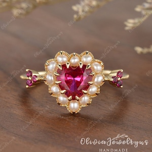 Peut inclure: Une bague en forme de cœur avec une grande pierre rouge sertie d'un halo de perles blanches. La bague a un délicat motif de flèche sur la bande.