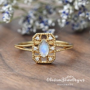 May include: A gold ring with a marquise-shaped moonstone surrounded by small diamonds. The ring has a delicate, intricate design and is set in a rectangular frame. The ring is handmade and is a beautiful piece of jewelry.