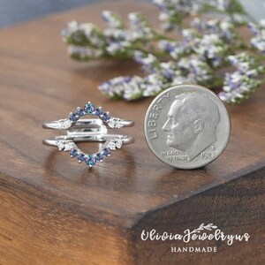Op de afbeelding: Een zilveren ring met een gesplitst ontwerp, met een rij kleine blauwe edelstenen en een rij kleine heldere edelstenen. De ring wordt getoond naast een Amerikaanse dime voor een vergelijking van de grootte.