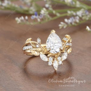 May include: A pear-shaped diamond engagement ring set with a delicate floral design. The ring features a gold band with a pear-shaped diamond in the center, surrounded by smaller diamonds and leaves. The ring is set on a wooden surface.