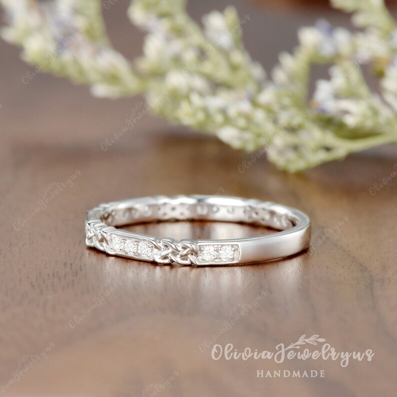 May include: A delicate silver band with a braided design and a row of small diamonds. The ring is on a wooden surface.