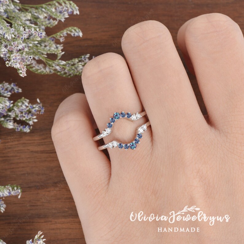 Op de afbeelding: Een delicate zilveren ring met een gebogen ontwerp met een rij blauwe edelstenen en kleinere witte edelstenen. De ring wordt aan een vinger gedragen.