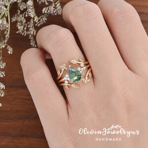 May include: A gold ring with a pear-shaped green gemstone surrounded by smaller diamonds. The ring features a delicate design of leaves and branches.