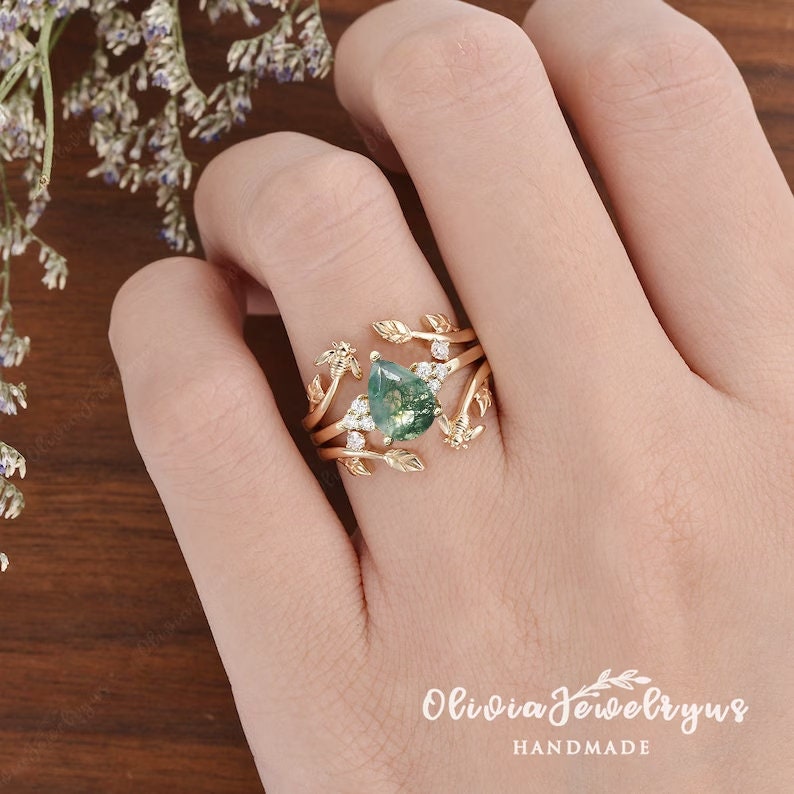 May include: A gold ring with a pear-shaped green gemstone surrounded by smaller diamonds. The ring features a delicate design of leaves and branches.