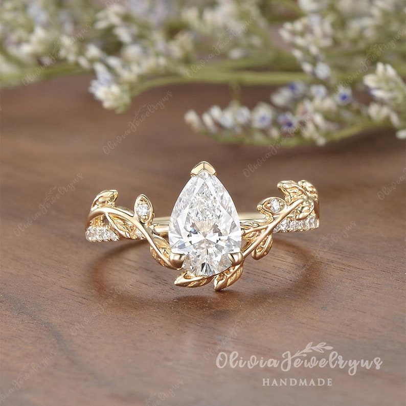 May include: A pear-shaped diamond engagement ring with a delicate gold band and leaf-shaped accents. The ring is set on a wooden surface.