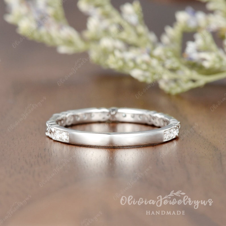 May include: A thin silver band with a row of small diamonds set in a delicate design. The band is on a wooden surface.