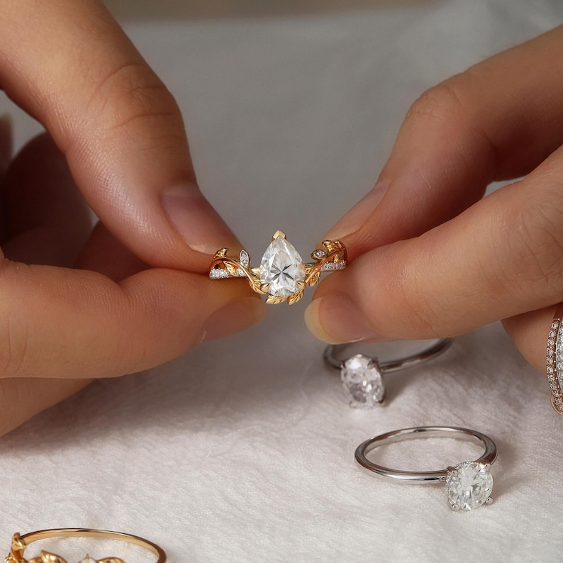 May include: A pear-shaped diamond engagement ring with a delicate gold band and leaf-like details. The ring is set with smaller diamonds on the band and leaves.