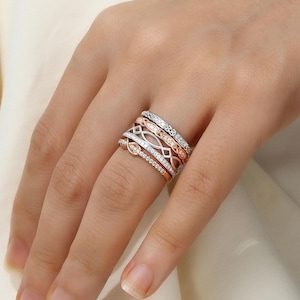 May include: A set of five delicate diamond rings stacked on a finger. The rings are made of white and rose gold and feature intricate designs.