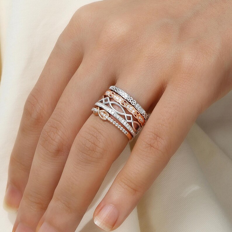 May include: A set of five delicate diamond rings stacked on a finger. The rings are made of white and rose gold and feature intricate designs.