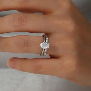 May include: A close-up of a hand wearing a silver oval engagement ring with a smaller band set with small diamonds on the finger.