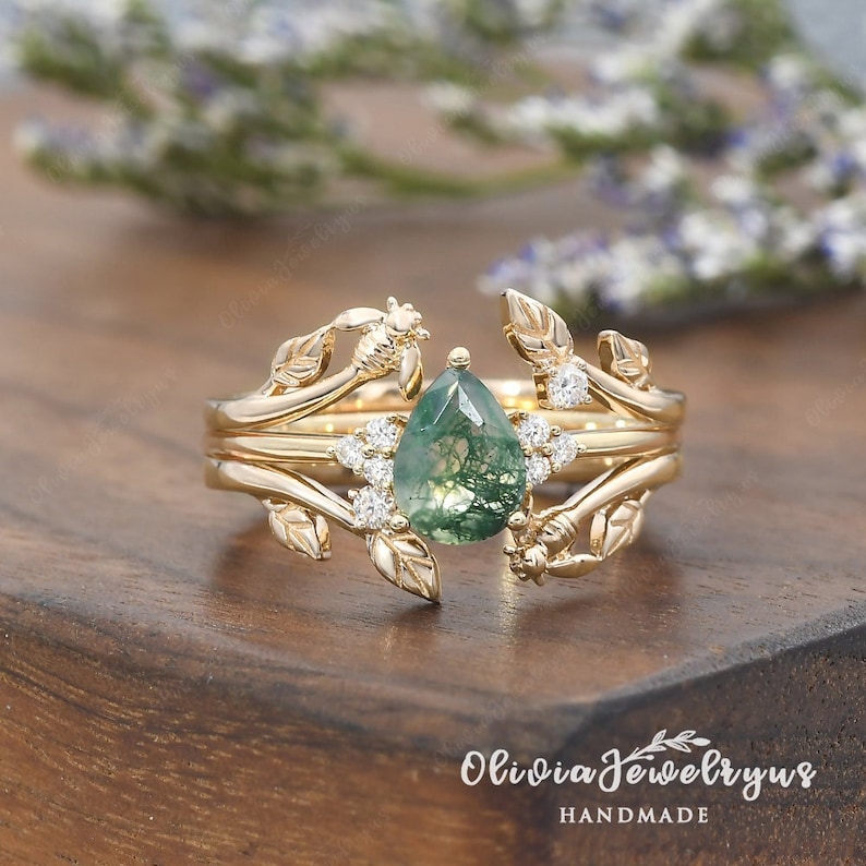May include: A gold ring with a pear-shaped green moss agate gemstone surrounded by small diamonds. The ring features a delicate design of leaves and bees.