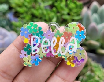 Puede incluir: Una etiqueta para perro de hueso de plástico transparente con un diseño floral colorido y el nombre "Bella" escrito en blanco.