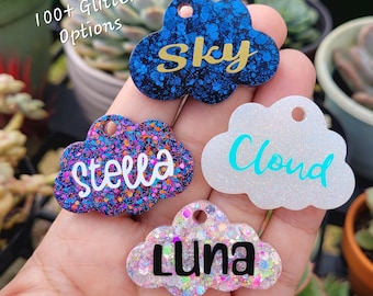 Può includere: Quattro etichette per nomi a forma di nuvola colorate con glitter. I nomi "Sky", "Stella", "Cloud" e "Luna" sono scritti sulle etichette.