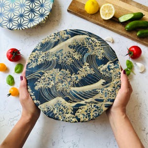 Peut inclure: Un film alimentaire réutilisable rond, bleu marine et or, avec un motif de vagues, tenu par des mains. D'autres films avec des motifs géométriques sont visibles, ainsi que des produits frais.