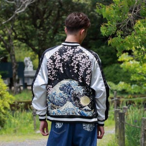 May include: A black and white satin bomber jacket featuring intricate embroidery on the back. The design displays a large blue ocean wave, a golden sun, a mountain peak, and pink cherry blossom branches against a black background.