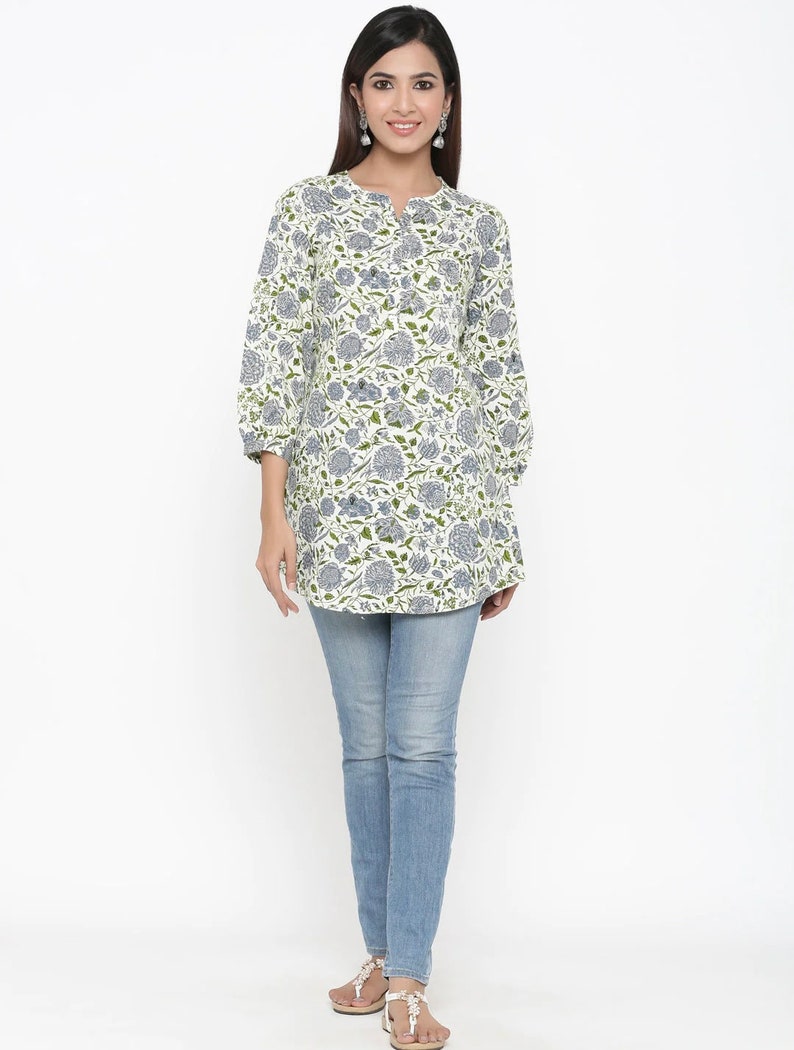 May include: A white cotton tunic top with a blue and green floral print. The top has a round neckline and three-quarter sleeves. The model is wearing the top with blue jeans.