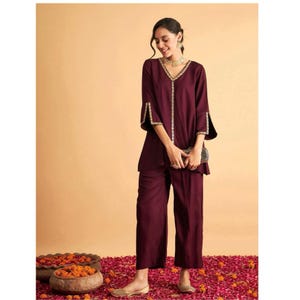 May include: A burgundy two-piece outfit with a V-neck top, gold trim, and flared sleeves, paired with wide-leg trousers. The outfit includes a small clutch and gold jewellery. This ensemble is suitable for special occasions. The outfit is a rich, deep colour.