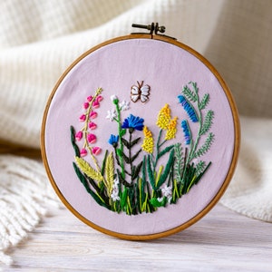 Può includere: Un telaio da ricamo cucito a mano con un design di prato di fiori selvatici colorati. Il design include fiori rosa, blu e gialli, fogliame verde e una piccola farfalla, il tutto su un tessuto lilla chiaro. Il telaio in legno è visibile.