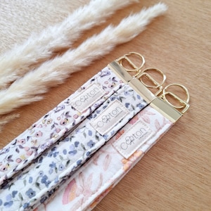 Peut inclure: Trois porte-clés en tissu à motifs floraux avec des attaches dorées. Les porte-clés présentent différents motifs floraux dans les tons de rose, de violet et d'orange. Chaque porte-clés porte l'étiquette "Coton Creations".