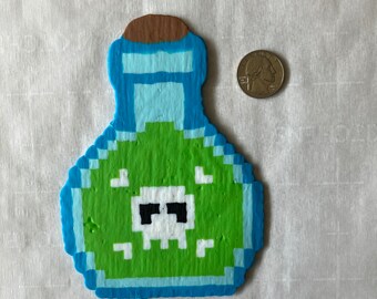 May include: A pixel art style potion bottle cutout featuring a green liquid center with a white skull icon, a blue border, and a brown cork top. A United States quarter coin is placed next to the object for scale, showing its relative size.