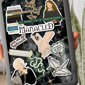 K&ouml;nnte beinhalten: Ein schwarzer Laptop mit verschiedenen Aufklebern. Die Aufkleber zeigen Illustrationen von H&auml;nden, Schlangen, Origami-Kranichen und Text. Der Text beinhaltet "A rose in a graveyard", "Manacled", "You are not replaceable", "I'm tired of not getting to choose you", "Be careful", "Don't die" und "Mudblood".