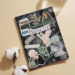 K&ouml;nnte beinhalten: Ein schwarzes Notizbuch mit verschiedenen Aufklebern. Die Aufkleber zeigen Illustrationen einer Frau, einer Schlange, einer Hand, die eine andere Hand h&auml;lt, eines Papierkranichs und eines B&uuml;cherstapels. Der Text auf den Aufklebern lautet "A Rose in a Graveyard", "Manacled", "You are not replaceable", "I'm tired of not getting to choose you", "Be Careful", "Don't Die", und "Only because you asked".