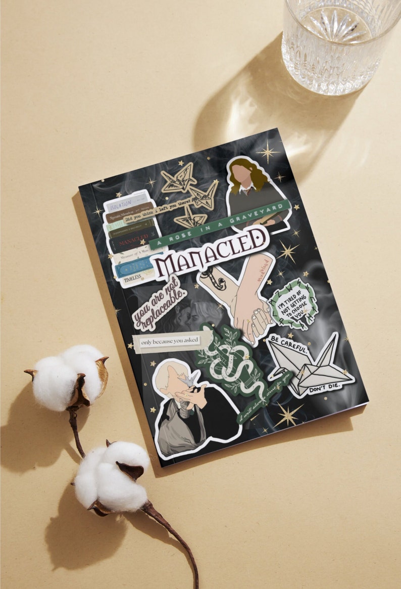 K&ouml;nnte beinhalten: Ein schwarzes Notizbuch mit verschiedenen Aufklebern. Die Aufkleber zeigen Illustrationen einer Frau, einer Schlange, einer Hand, die eine andere Hand h&auml;lt, eines Papierkranichs und eines B&uuml;cherstapels. Der Text auf den Aufklebern lautet "A Rose in a Graveyard", "Manacled", "You are not replaceable", "I'm tired of not getting to choose you", "Be Careful", "Don't Die", und "Only because you asked".