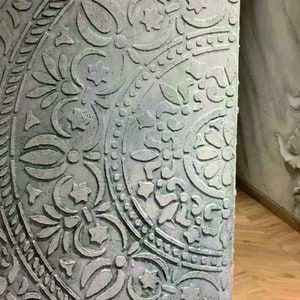 Könnte beinhalten: Nahaufnahme eines Wandabschnitts mit einem detaillierten, geprägten Blumen- und geometrischen Muster. Die Wand hat ein verwittertes, gealtertes Aussehen mit einer gedämpften Grün- und Graupalette. Das Design umfasst kreisförmige und sternförmige Elemente.