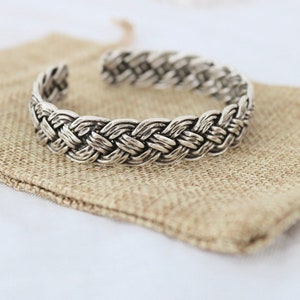 Könnte beinhalten: Ein silbernes geflochtenes Armreif mit einem gewebten Design. Das Armband liegt auf einem braunen Jute-Stoffhintergrund.