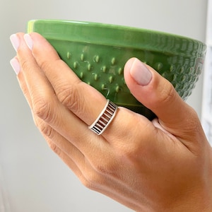 Könnte beinhalten: Ein silberner Ring mit geometrischem Design. Der Ring wird an einem Finger getragen. Die Person hält eine grüne Keramikschüssel.