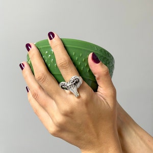 May include: A silver ring featuring a ram's head design. The ring is worn on a finger, and the person is holding a green bowl.