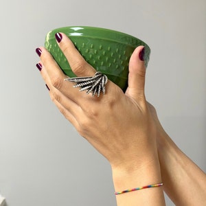 Könnte beinhalten: Ein silberner Ring mit einem großen, komplizierten Design, das an eine stilisierte Feder oder einen Flügel erinnert. Der Ring wird am Zeigefinger einer Hand getragen, die eine grüne Keramikschale hält.